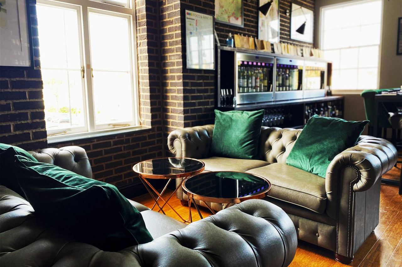 The Wine Library upstairs at Tivoli Wines: deep-green Chesterfield sofas, copper coffee tables, exposed-brick wall, with the Enomatic wine dispensers stocked along the back wall